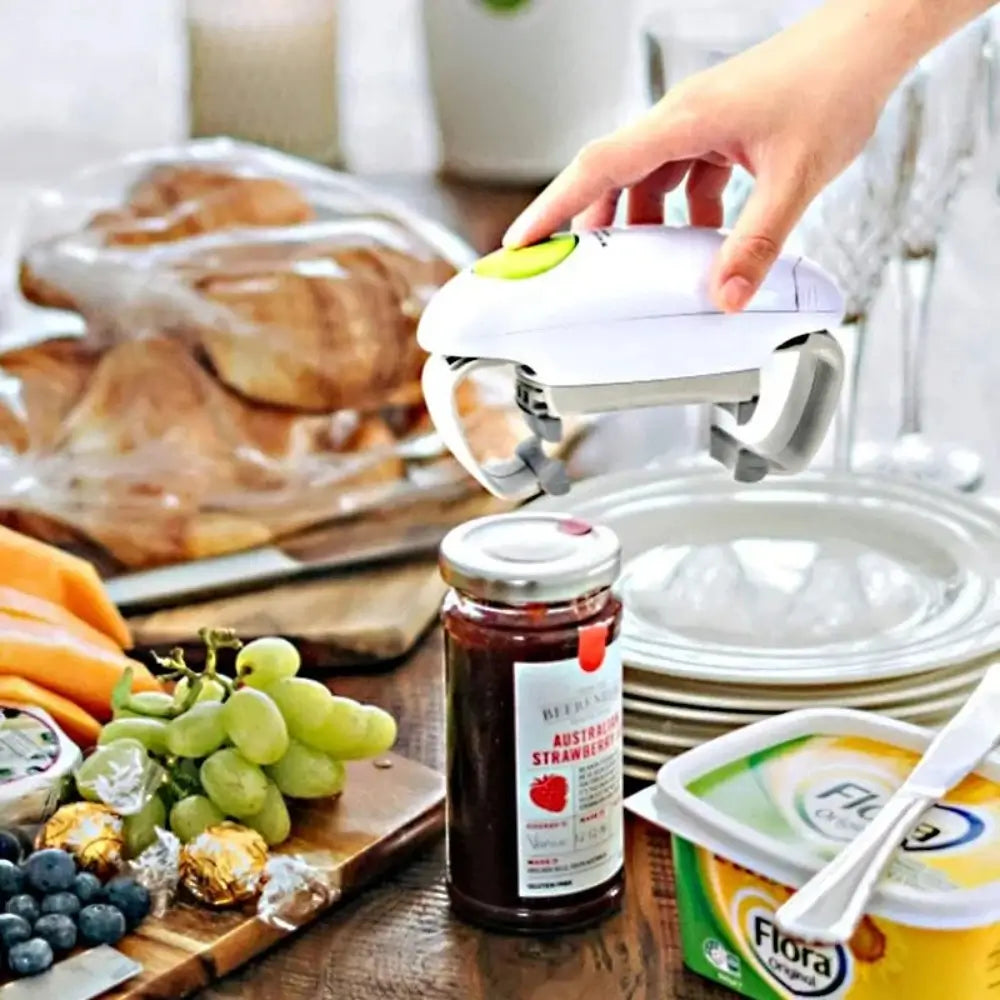 The automatic electric jar opener about to be placed on a strawberry jam jar which is placed on a table surrounded by fruits, breads, a tub of butter and a stack of plates.