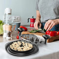 The chicken breast shredder in black resting on a bench top in a kitchen. A women chopping various vegetables on a chopping board, a bowl of salad and salt and pepper shakers to the left, with other various kitchen appliances and utensils in the background.