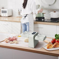 The dumpling maker machine on a kitchen bench surrounded by food, condiments and kitchen utensils. A women in the kitchen in the background tending to a cooking pot.